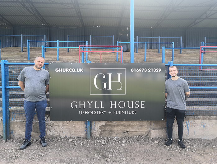 Joe McAlpine and Ethan Black in front of the Ghyll House Upholstery's advertising board at Workington Town’s Derwent Park stadium.